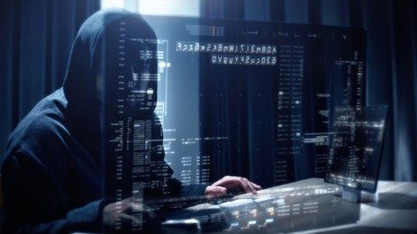 A threat actor typing on a keyboard with screens in front displaying lines of code, evoking the theme of social engineering or cybersecurity.